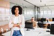 © Vadim Pastuh - A confident African-American woman with a radiant smile stands with her arms crossed. The background reveals a coworker focused on his screen, highlighting a professional and dynamic work environment