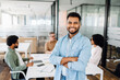 © Vadim Pastuh - A confident young Indian male office employee in blue stands with his arms crossed in the foreground, with his team in discussion behind him, signaling a modern and proactive work environment