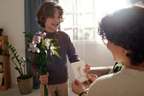Fototapeta  - Happy cute boy with bunch of flowers passing handmade greeting card to his mother while congratulating her on traditional spring holiday