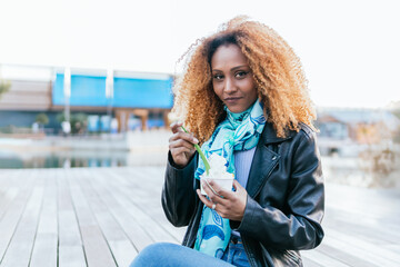 Canvas Print - A woman with curly hair is sitting on a wooden bench and eating ice cream. She is wearing a black jacket and a blue scarf. The scene is casual and relaxed, on the outdoors of shopping mall. She's look