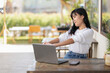 © Wasana - Relaxed Asian woman stretching her arms while taking a break from work using a laptop at an outdoor cafe.