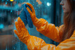 Close up of woman hands in orange gloves cleaning the window with blue rag, microfiber cloth . Hand wiping dirty glass on the blue city street background. Banner