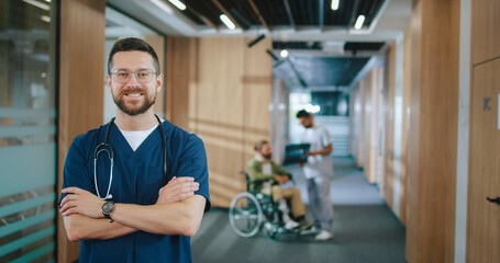 Portrait of attractive successful Caucasian doctor in uniform with stethoscope looking at camera and smiling. Happy professional medical worker standing at corridor with patient on background.