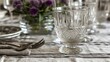 a close up of a glass on a table with silverware and a vase with purple flowers in the background.