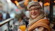 A smiling elderly woman in a coat, scarf and hat sitting with glass of orange juice in her hand at street cafe