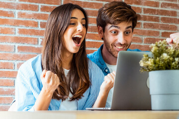 One young adult couple in front of a laptop with winner excited joyful expression and gesture together. Man and woman and online business job success concept small business lifestyle at home leisure