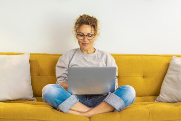 One woman smiling and using laptop computer at home sitting comfortably on a yellow sofa in living room. Smart working female people notebook. Surfing the web. Enjoying technology and connection