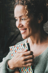 Wall Mural - Happy smiling expression woman hold book and look outside the window at home. Dark background. Joyful face portrait of female people in indoor leisure activity alone. Attractive middle age lady indoor