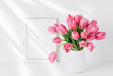 Fototapeta Tulipany - White frame mockup and pink tulips in a white vase on a white table by the wall