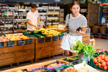 Positive young female shopper making purchases in supermarket, looking with interest for ripe organic courgettes