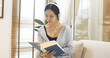Happy asian woman use headphones listen to music reading book sitting on cozy sofa in living room at home. Female asian people work at home research book read magazine smiling happy reader lifestyle