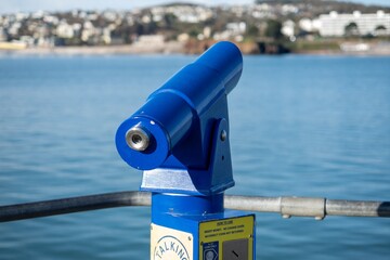 A blue viewing telescope at a seaside location looks across a calm blue sea to land in the distance at a popular holiday location