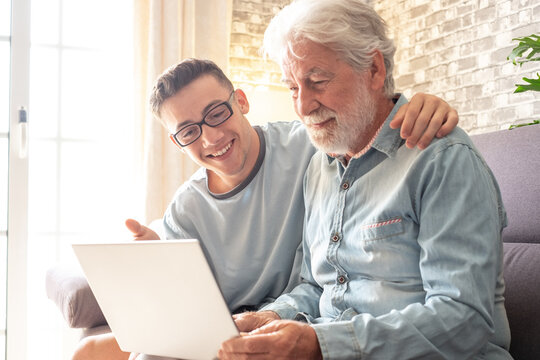 Smiling experienced young boy with laptop teaching and showing new computer technology to his old grandfather for surfing internet, shopping online, managing bank. Old man learns to use the computer