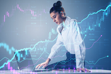 Wall Mural - Attractive young businesswoman using laptop while sitting on desk with creative glowing candlestick forex chart on blurry background. Financial trade and market concept. Double exposure.