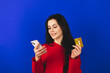 © zvkate - Online shopping. Portrait of smiling brunette curly hair woman paying with plastic credit card on smartphone app, standing with mobile phone and bank card against blue background. Happy emotions.