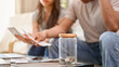 © bongkarn - A jar of coins on a coffee table with a stressed couple planning their finances.