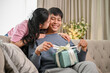 © bongkarn - A lovely Asian wife is kissing her husband's cheek on a couch after surprising him with a present.
