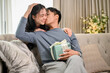 © bongkarn - A romantic Asian husband is kissing his wife's cheek on a couch to thank her for a special present.