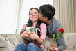 © bongkarn - A romantic Asian husband kisses his wife's cheek after surprising her with a present and flowers.