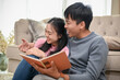 © bongkarn - A cheerful young Asian couple is enjoying reading a book together in the living room.