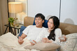 © bongkarn - A happy young Asian couple in pajamas is enjoying watching television or a movie together in bed.