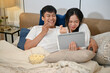 © bongkarn - A happy Asian couple are enjoying watching a movie on a tablet and eating popcorn in bed together.
