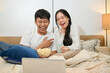 © bongkarn - A cheerful Asian couple is laughing, enjoying watching a movie on a laptop in bed together.
