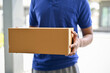 © bongkarn - A cropped shot of a delivery man in a blue shirt holding a cardboard box in front of the door.