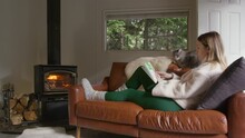 People On Weekend Getaway In Remote Destination. Relaxed Woman Sitting With Grey Cat On Comfortable Leather Coach. Female Reading Interesting Book, Warming At Wood Burning Stove In Cozy Forest Cabin