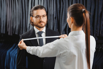 Wall Mural - Woman tailor taking measurements of young handsome man, fitting bespoke suit to model. Lux Atelier Classic Menswear