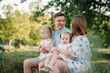 © Serhii - A loving couple with two childs. Mother, father embrace kids and spend time together. A happy family is sitting on bench on shore of lake at sunset. Mom, dad hug children daughter in the summer park.