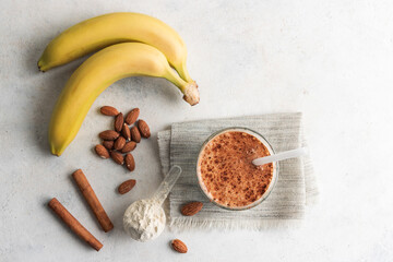 Wall Mural - Glass with protein drink, healthy milkshake smoothie on white table with bananas, protein powder in measuring spoon, almond nuts and cinnamon sticks, top view