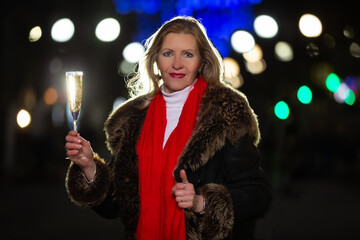 Wall Mural - Portrait of a beautiful woman with a glass of champagne in an illuminated evening city. Happy evening celebration moment.