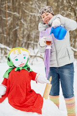 Wall Mural - Woman pours hot beverage from jug into glass standing near stuffed Maslenitsa in winter park