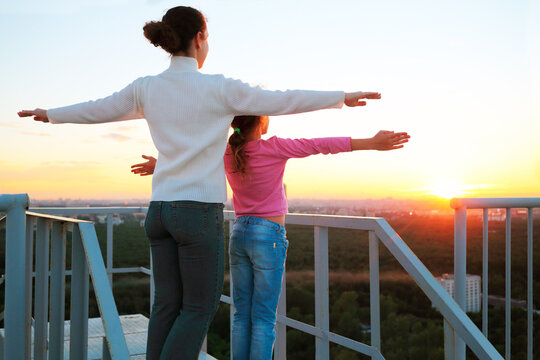 Mother and daughter standing with arms outstretched to sides on roof of multistory building in evening and look at sunset