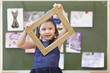 © Pavel Losevsky - Girl looking through decorative frame on background of pictures hung on blackboard in classroom, looking at camera, turned frame diagonally