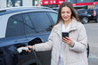 © Nomad Pixel - Young woman charging her electric car at a charging station in the city. Eco fuel concept. The concept of environmentally friendly transport. Recharging battery from charging station.
