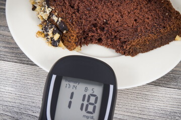 Glucose meter with result sugar level and sweet chocolate cake. Measuring and checking sugar level during diabetes