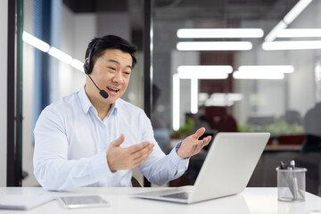 Canvas Print - Smiling asian man in white shirt working in office at desk, wearing headset, talking on video call through laptop gesturing with hands.