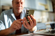 © Marko Geber - Close-up of a man using a smartphone with a laptop and documents on the table