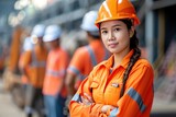 Fototapeta Przestrzenne - An asian worker in orange safety gear and helmet stands confidently, arms crossed, with colleagues in similar attire blurred in the background, suggesting teamwork at an industrial site.