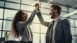 © MP Studio - A man and a woman in business attire are in an office setting, happily giving each other a high-five, suggesting a successful moment or celebration.