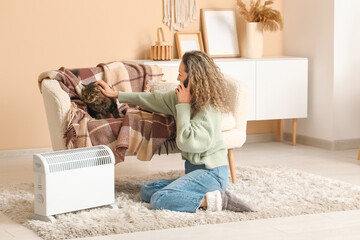 Sticker - Happy mature woman petting cute cat on sofa near electric heater at home