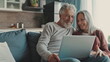 © YauheniyaA - Cheerful and happy middle-aged spouses spending time with a laptop at home. Handsome grey-haired man and beautiful good-looking woman sit on the sofa, using laptop and smile