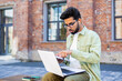 © Liubomir - Young indian man in olive shirt sitting on bench at campus with pc on laps and wired earphones. Concentrated student pointing with finger at laptop screen while talking with group mate in online call.