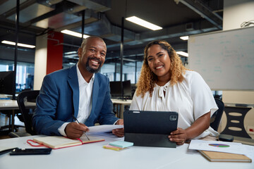 Two happy multiracial colleagues looking at camera and smiling next to digital tablet in office