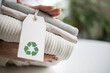 Woman's hands hold a stack of clean neat knitted clothes with a label with a recycling sign.