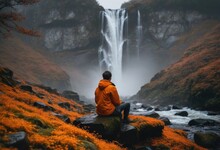 Person Taking A Break From Their Journey, Sitting On Wet Rocks With Patches Of Orange Moss, Admiring A Waterfall Obscured By Thick Fog.