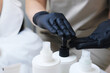 © ribalka yuli - Beautician Dispensing Cosmetic Product from Container. A beautician's hands, clad in black gloves, are pressing a pump to dispense a professional skincare product from a white bottle.