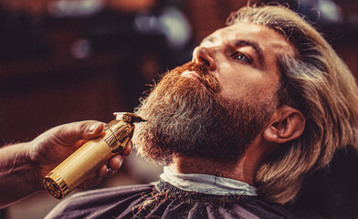 Wall Mural - Barber preparing hair clipper for bearded man, barbershop background. Barber works with a beard clipper. Hipster client getting haircut. Hands of a hairdresser with a beard clipper, closeup